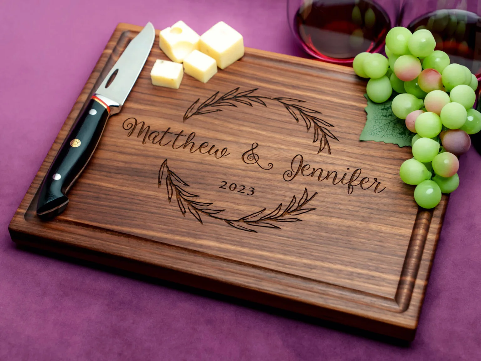 walnut charcuterie board with cheese knife green grapes cheeses and two glasses on red wine on purple background