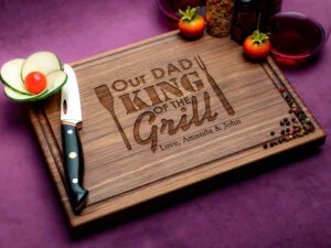 wooden cutting board with "Our Dad, King of the Grill" engraved knife with sliced cucumber and cherry tomato and peppers on a purple surface