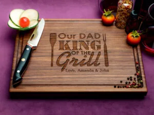 wooden cutting board with "Our Dad, King of the Grill" engraved surrounded by a knife, sliced vegetables, spices, and tomatoes.