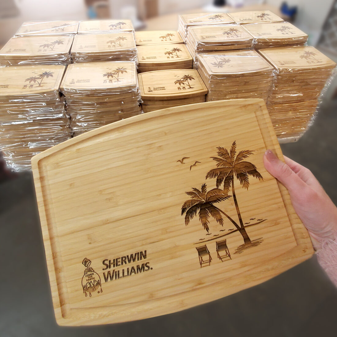 Corporate Gifts: 7 Reasons Why Personalized Cutting Boards Are the Best Choice 1 Personalized bamboo cutting board engraved with a tropical scene and the Sherwin Williams logo, crafted as a corporate gift. A stack of similar boards in the background, each wrapped and ready for distribution