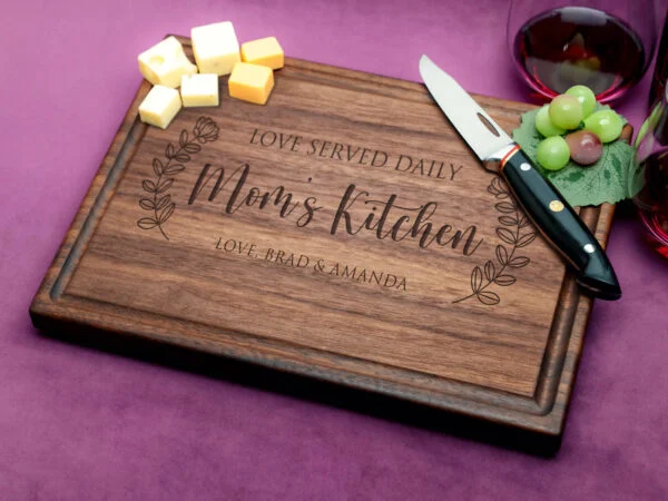 A walnut cutting board with a juice groove, elegantly engraved with 'Mom's Kitchen'. Highlighting durable wood craftsmanship, this personalized piece combines practicality and sentimentality for a perfect kitchen accessory.