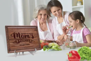 Handcrafted walnut cutting board adorned with 'Mom's Kitchen' design and personalized details. Encircling the board, a family joyfully prepares a meal, creating cherished memories in their bustling kitchen atmosphere.