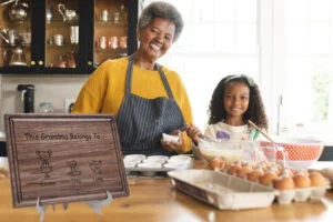 A personalized walnut cutting board with a family members design and customized engraving, resting on a kitchen counter. Nearby, a grandmother and her granddaughter joyfully bake together, surrounded by warmth and love in their kitchen.